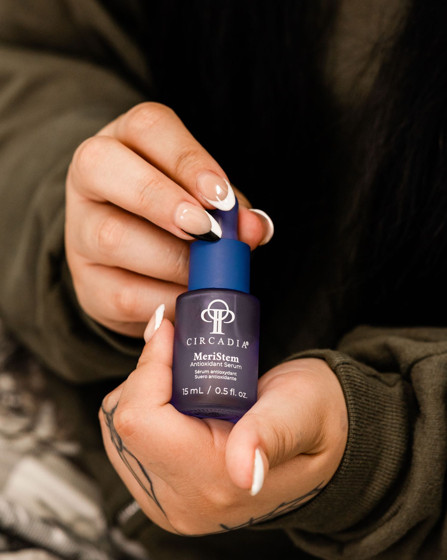 Tattooed hands holding a small blue Circadia MeriStem antioxidant serum bottle with French tip nails.