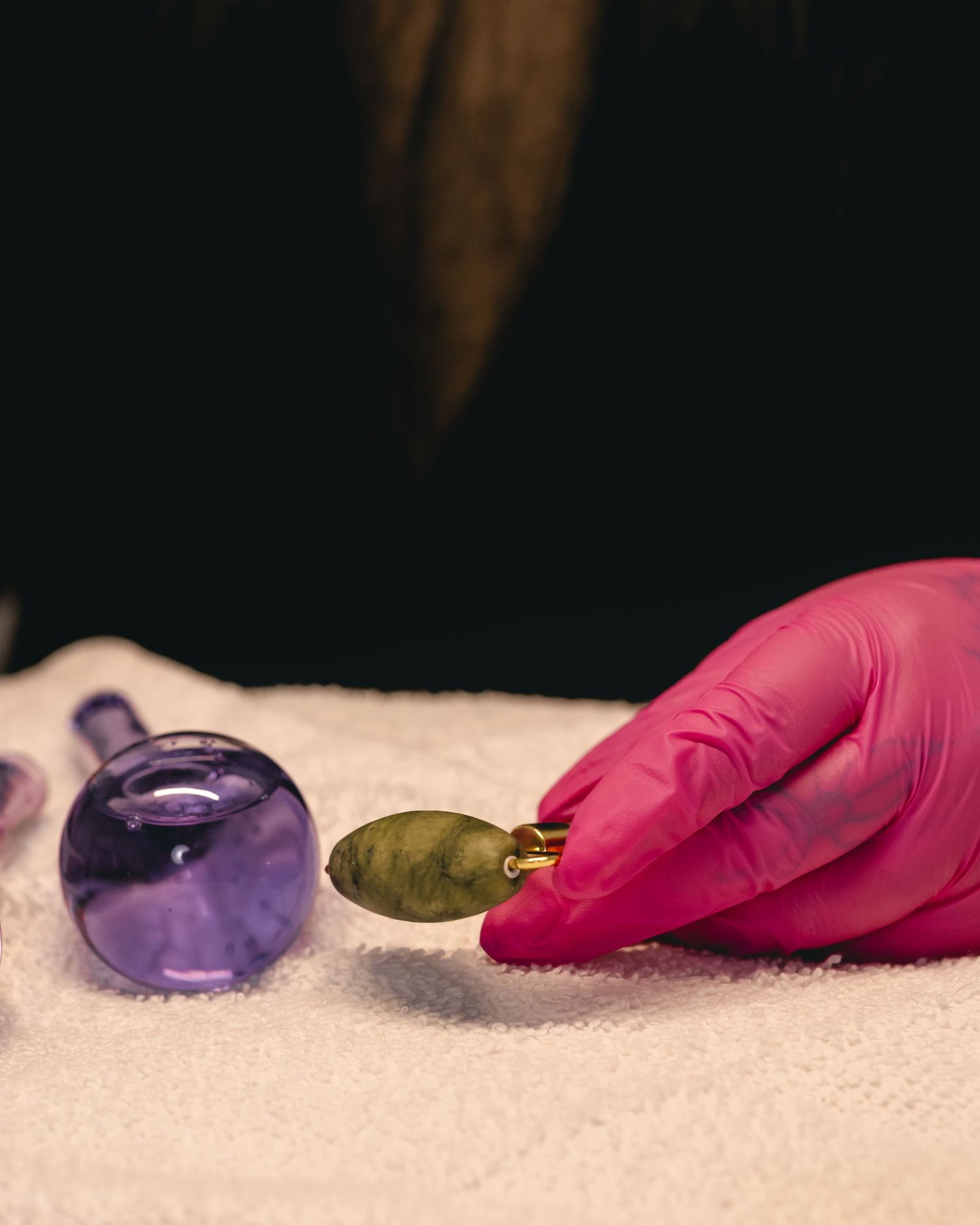 Gloved hand in pink latex holding a small green leaf near purple glass facial cupping tools on white towel.