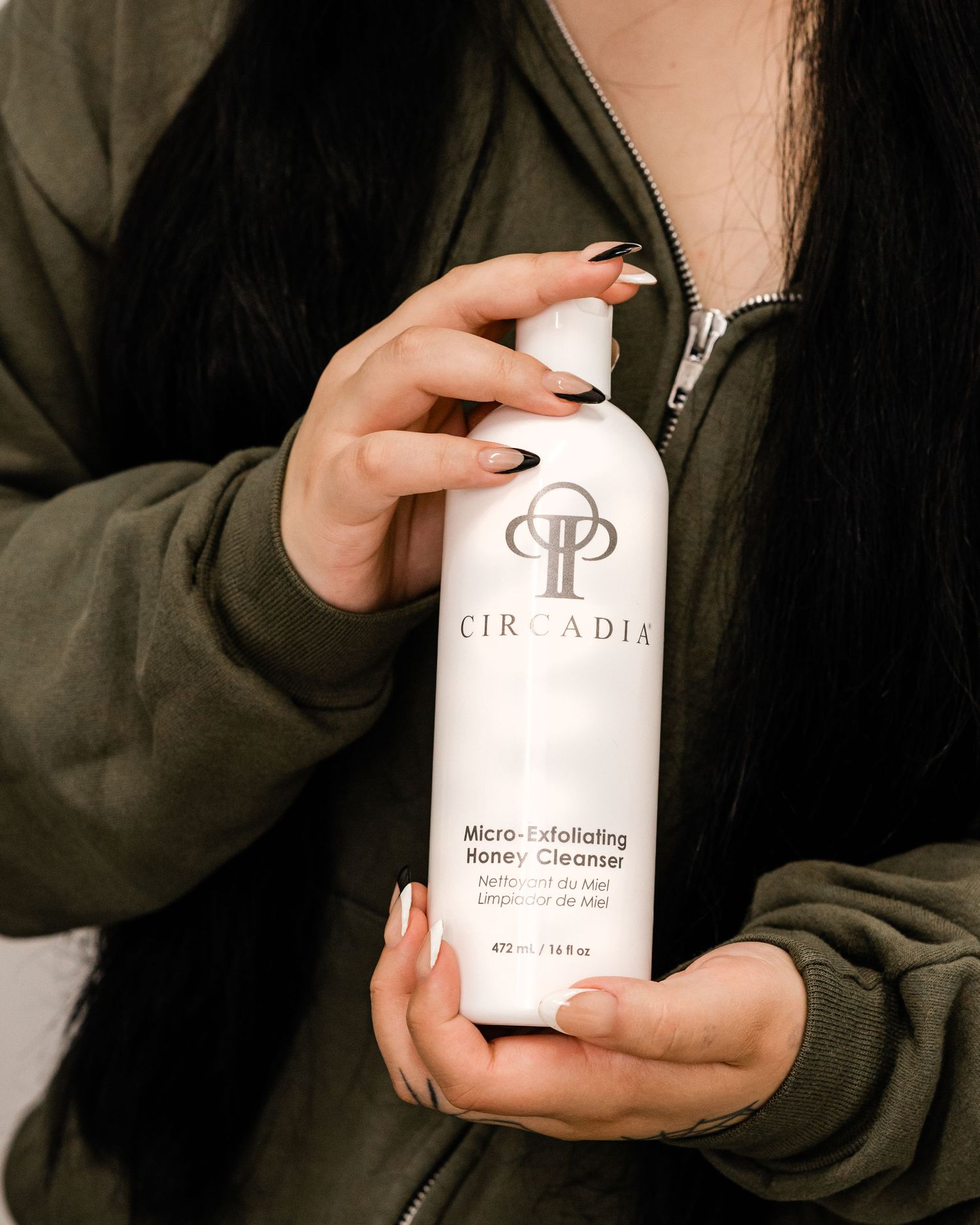 Woman in olive hoodie holding Circadia Micro-Exfoliating Honey Cleanser bottle with black manicured nails.