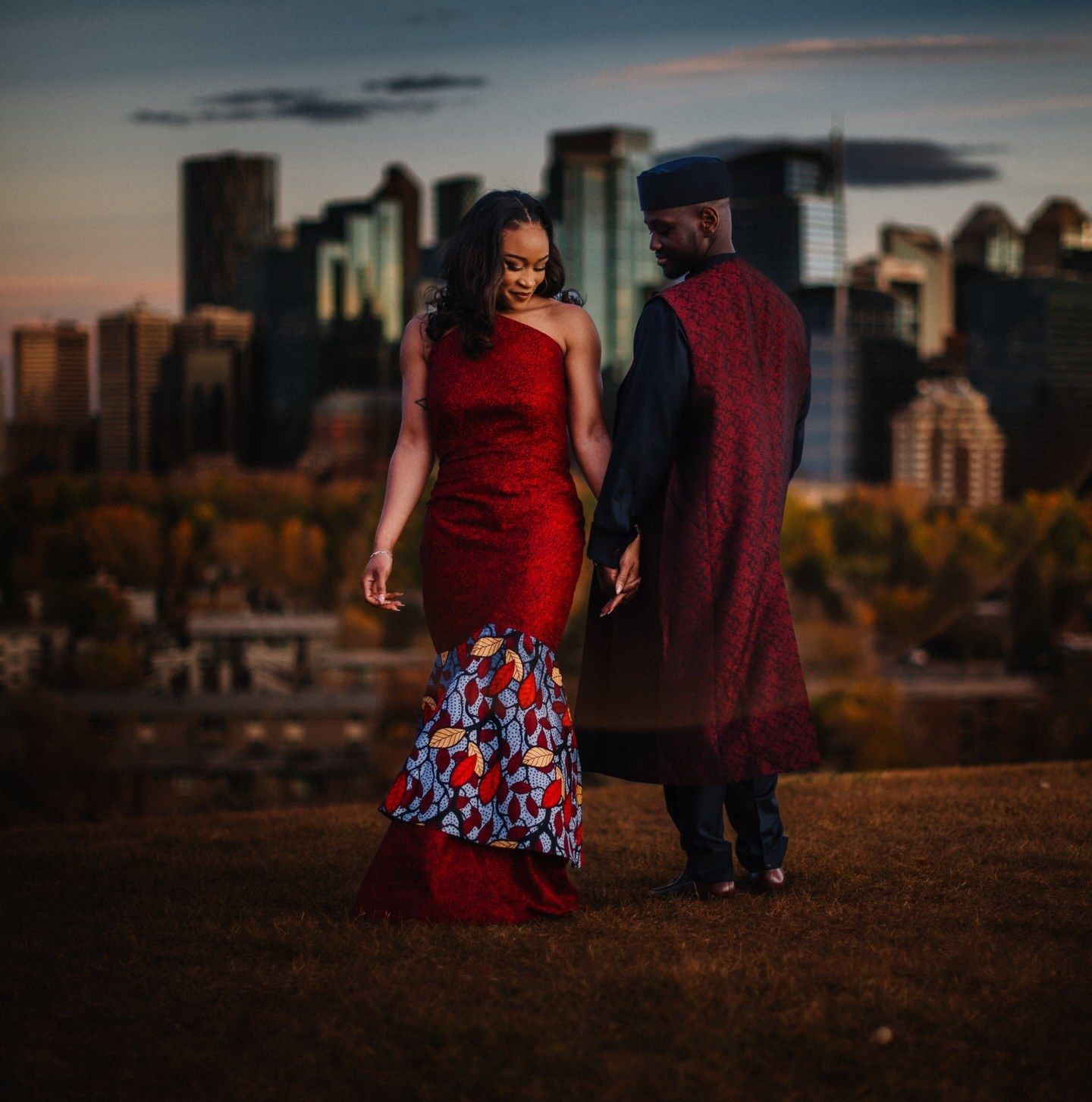 Couple in elegant red African-inspired attire holding hands on a grassy hill at sunset with a glowing city skyline behind them.