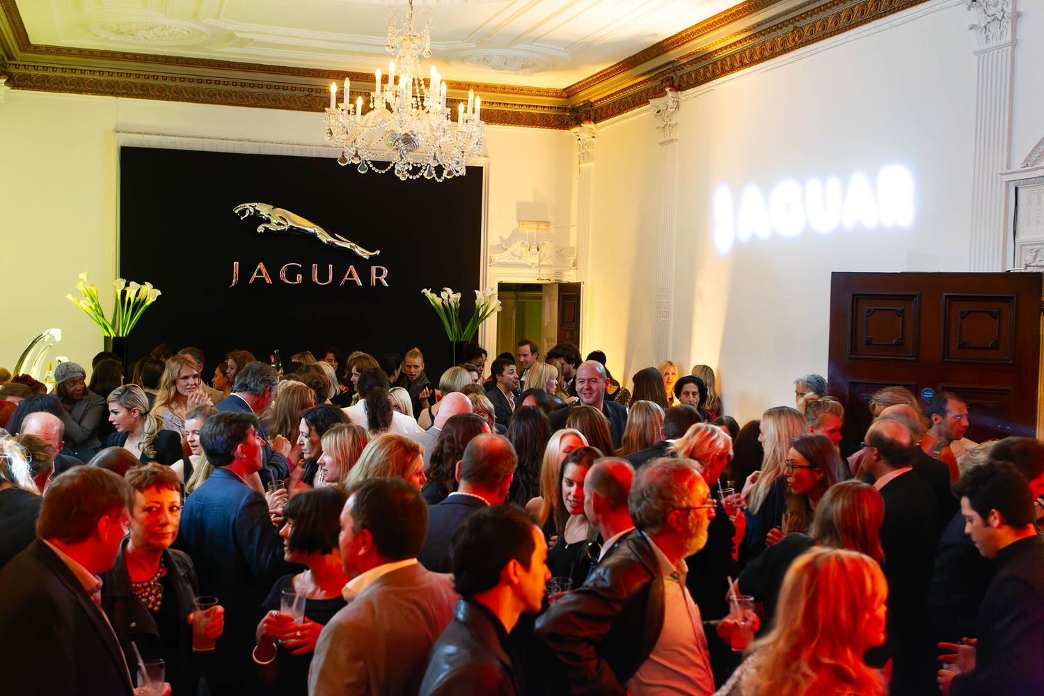 Crowded Jaguar event in a chandeliered reception room with the Jaguar logo on the wall