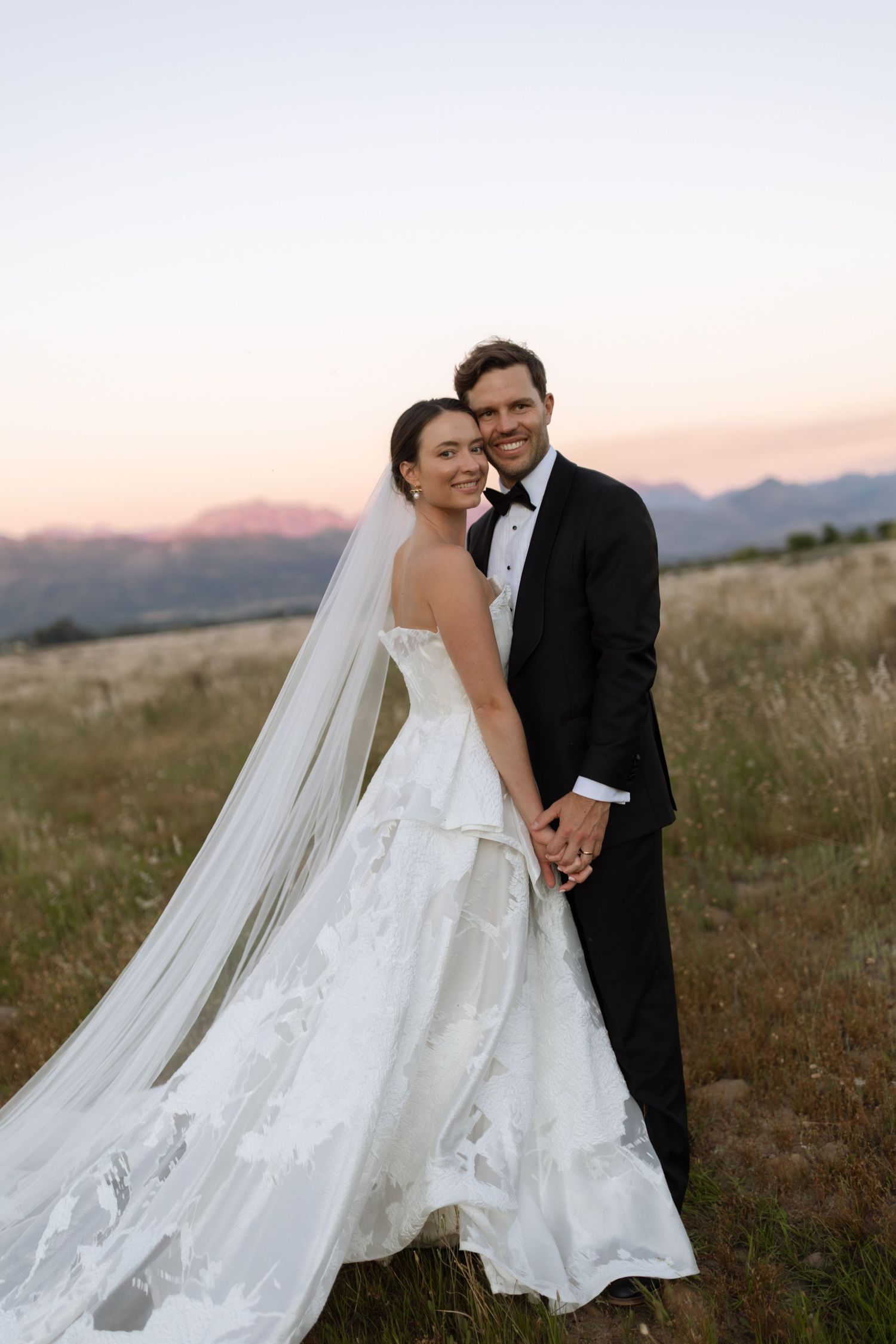 Bride in a strapless floral lace wedding gown and long veil embraces her groom in a black tuxedo in a field at sunset with mountains behind.
