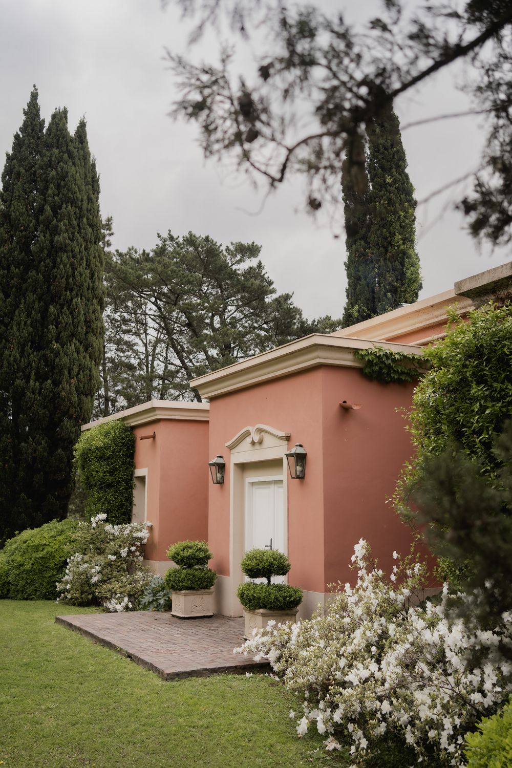Pink stucco villa with lantern sconces, topiary shrubs, and white flowering bushes beside a green lawn and tall cypress trees.