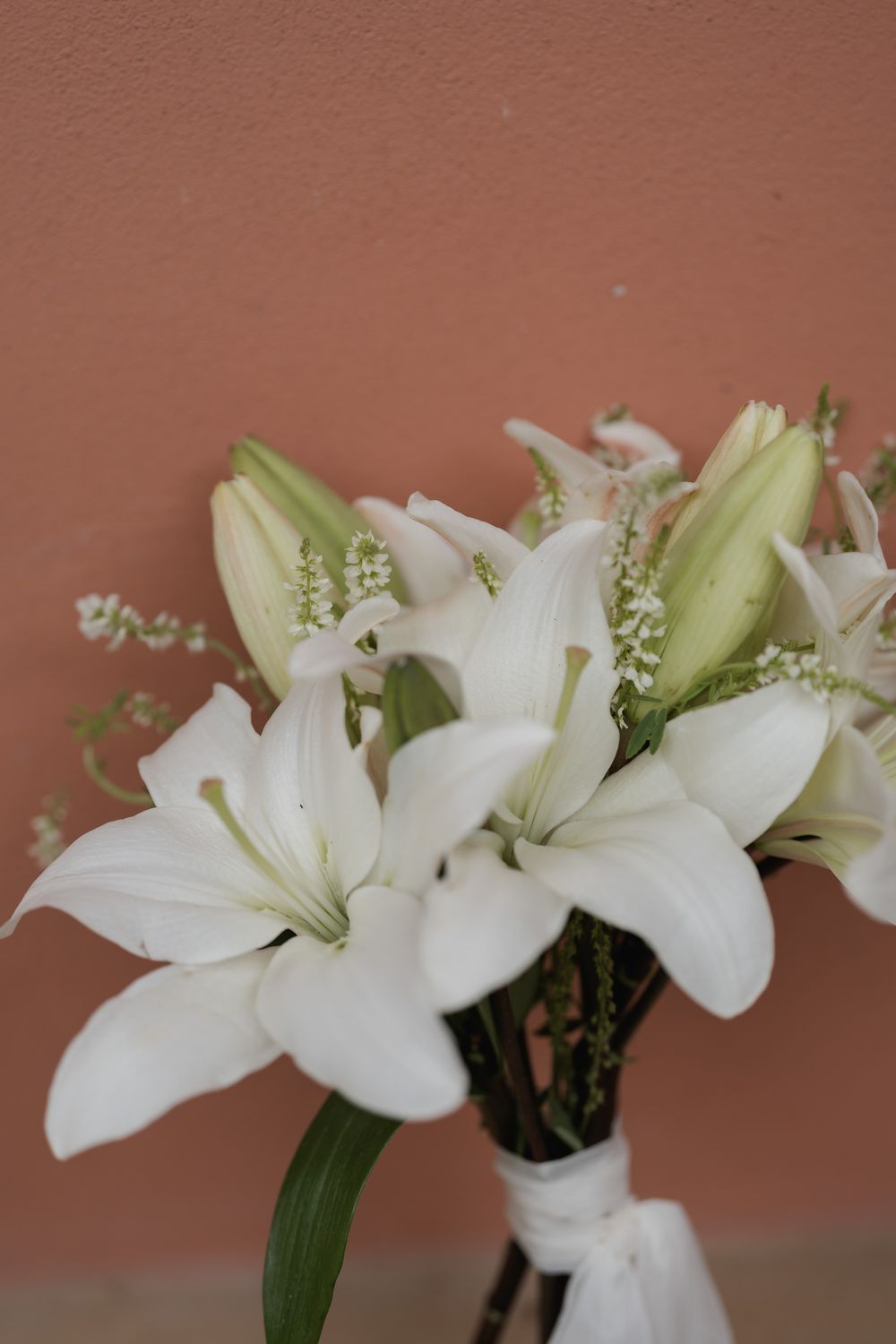 White lily bouquet with open blooms, green buds and delicate filler flowers tied with a white ribbon against a terracotta wall.