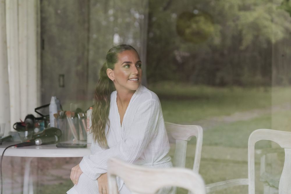 Smiling woman with long ponytail in a white robe sits on a white chair near a table with beauty supplies, with green trees visible outside.