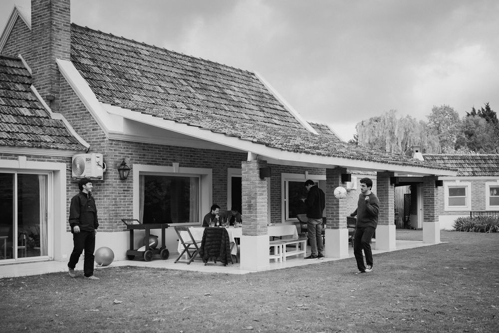 Black and white photo of several people standing on the covered porch and lawn of a brick house with a tiled roof under a cloudy sky.