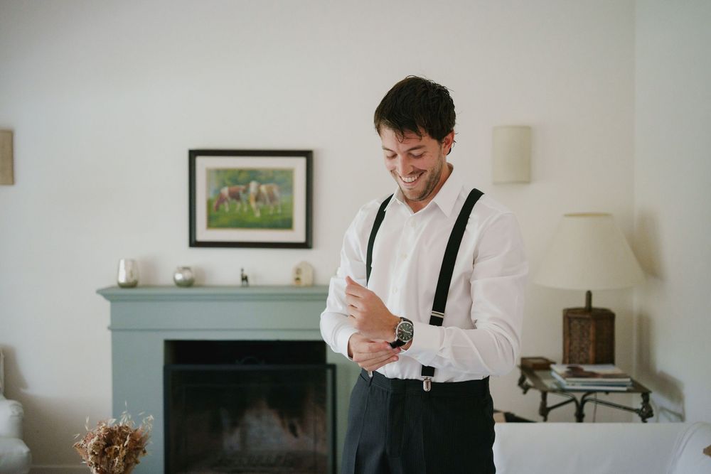 Smiling man in a white shirt and black suspenders adjusts his cuff and watch in a bright living room with a mint green fireplace.