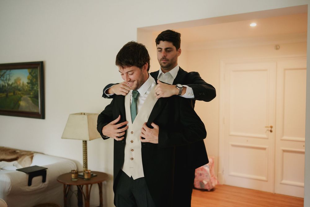 A groomsman helps a man in a vest and green tie put on his dark suit jacket in a bright room before the wedding.