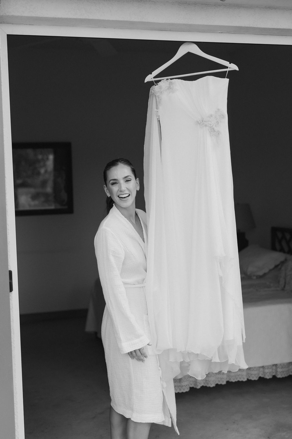 Smiling bride in a white robe stands in a doorway beside her strapless wedding gown hanging on a hanger, in black and white.