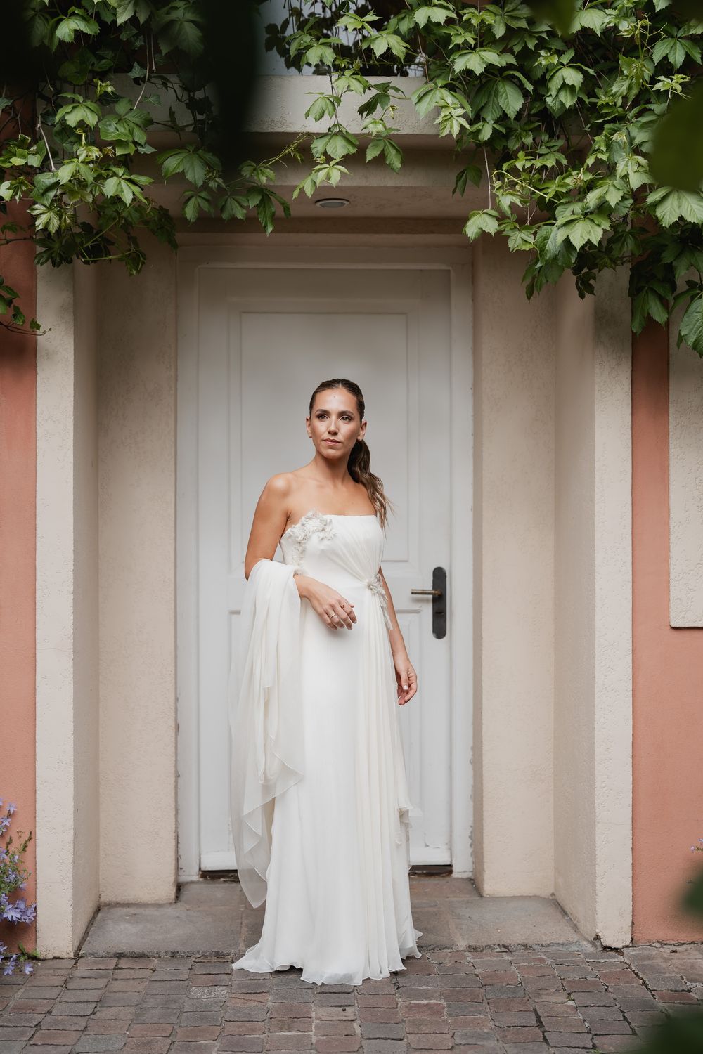 Bride in a flowing white strapless chiffon wedding gown with a floral detail stands before a white door framed by ivy and peach walls.