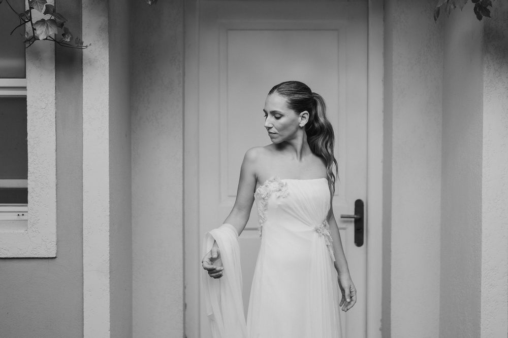 Black and white photo of a bride in a strapless wedding gown with her hair in a ponytail, holding a veil beside a white door.
