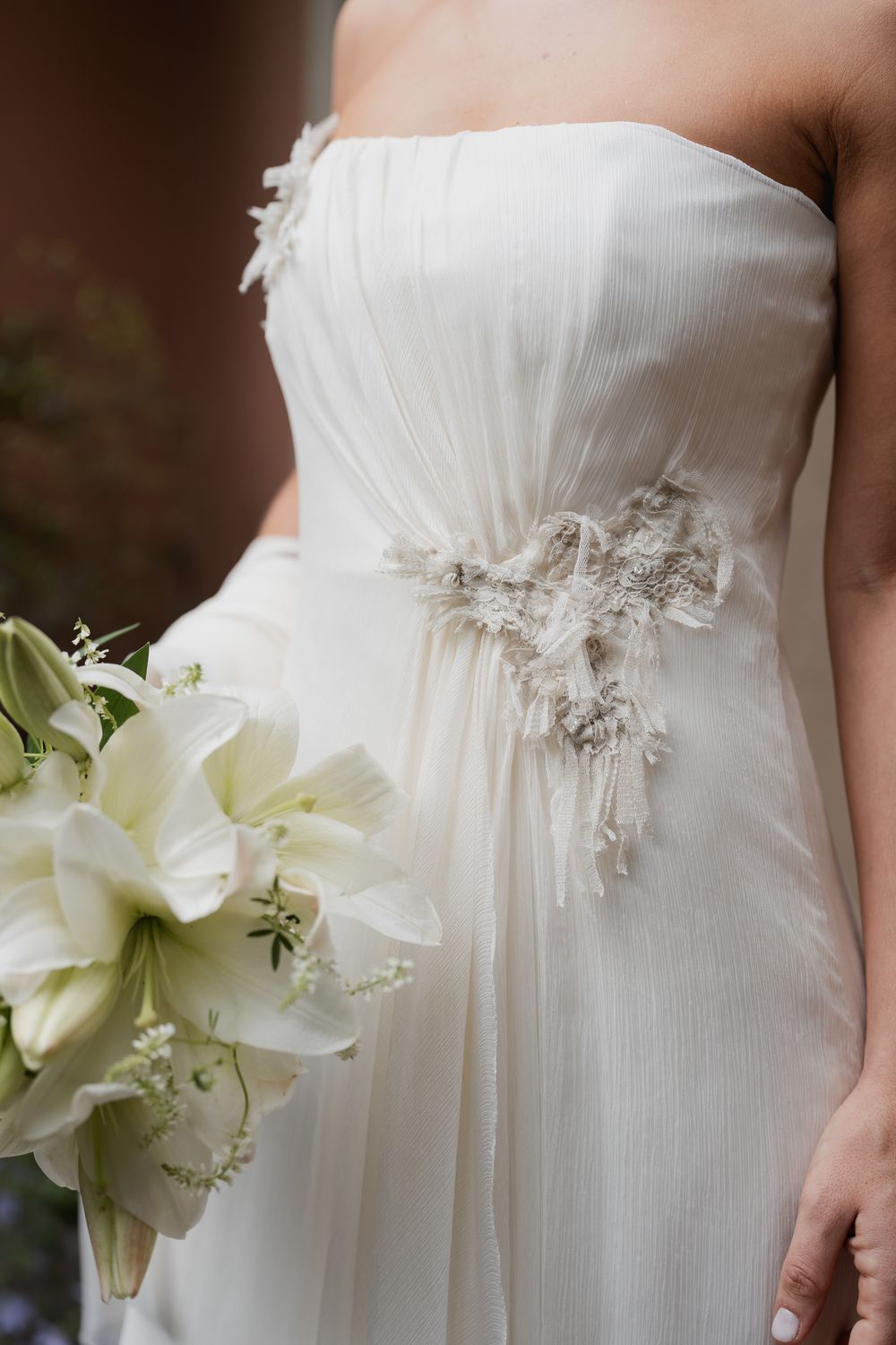 Bride in a strapless ivory chiffon wedding gown with beaded floral detail holding a bouquet of white lilies.