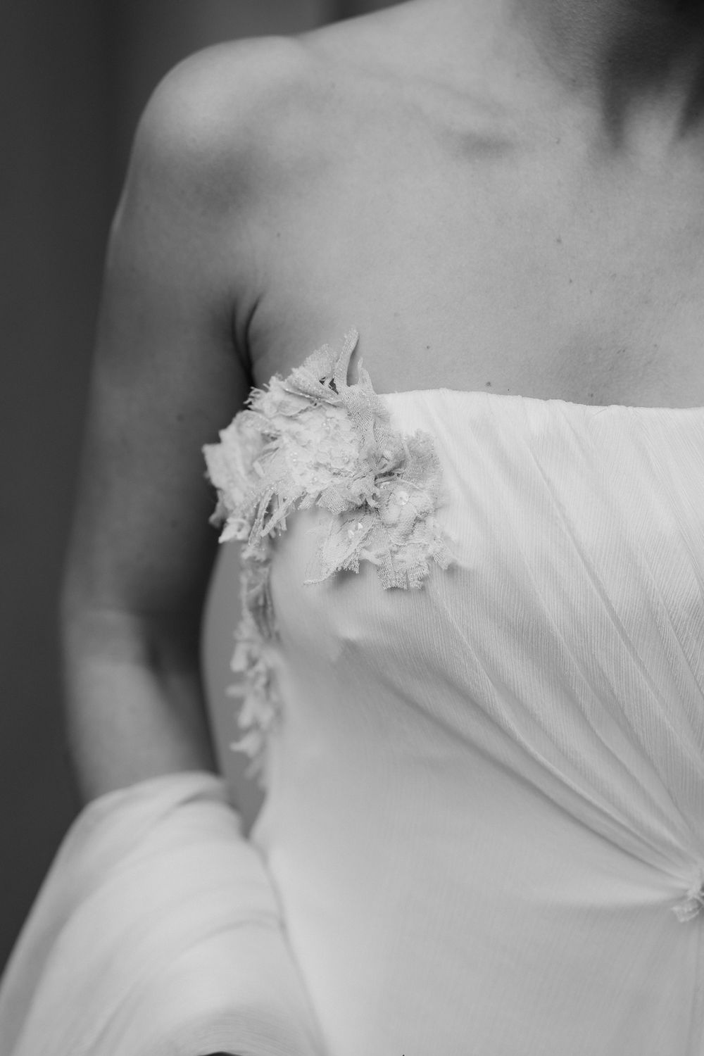 Black and white close-up of a strapless bridal gown with ruched bodice and delicate lace floral appliqué at the shoulder.
