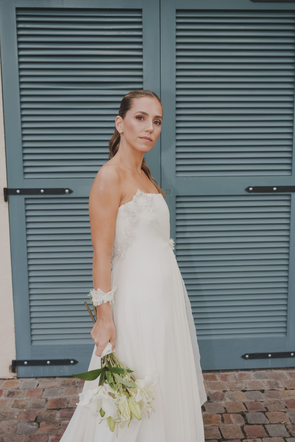 Bride in a strapless white lace gown holding a white floral bouquet stands before blue-gray shuttered doors on a brick patio.