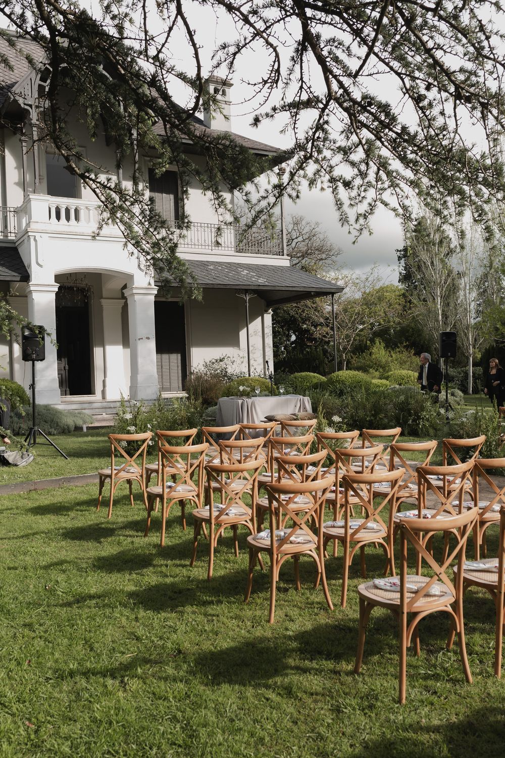 Wooden cross-back chairs arranged in rows on a green lawn for an outdoor wedding ceremony beside a historic white mansion.