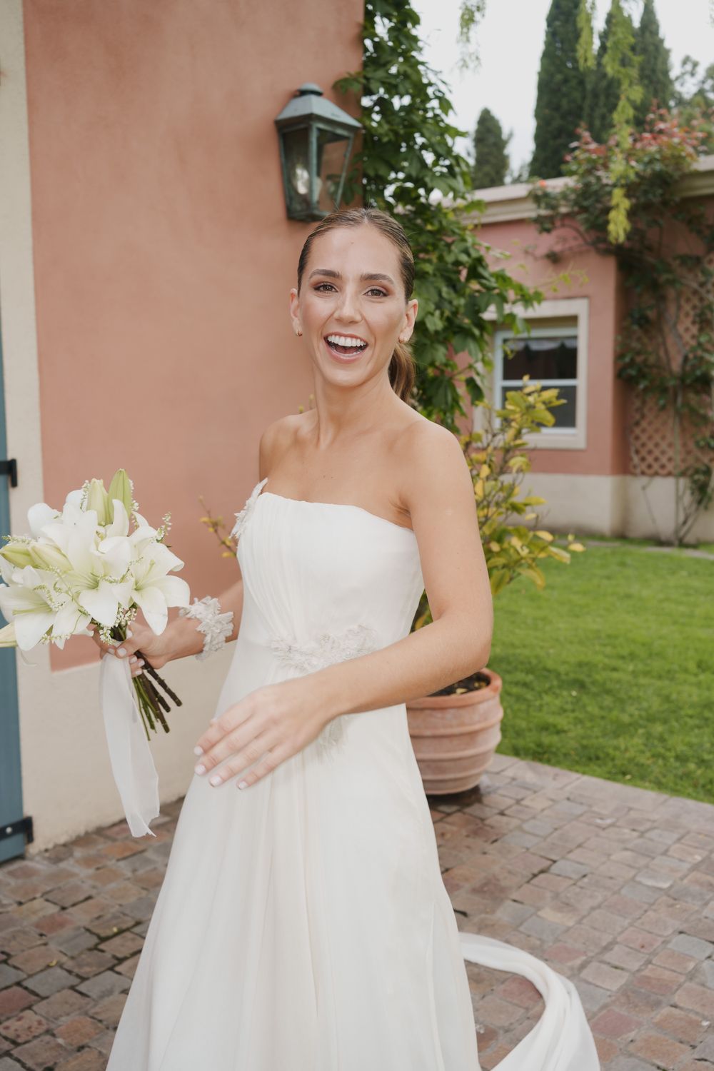 Smiling bride in a strapless white wedding gown holds a bouquet of white lilies on a brick patio beside a peach-colored building.