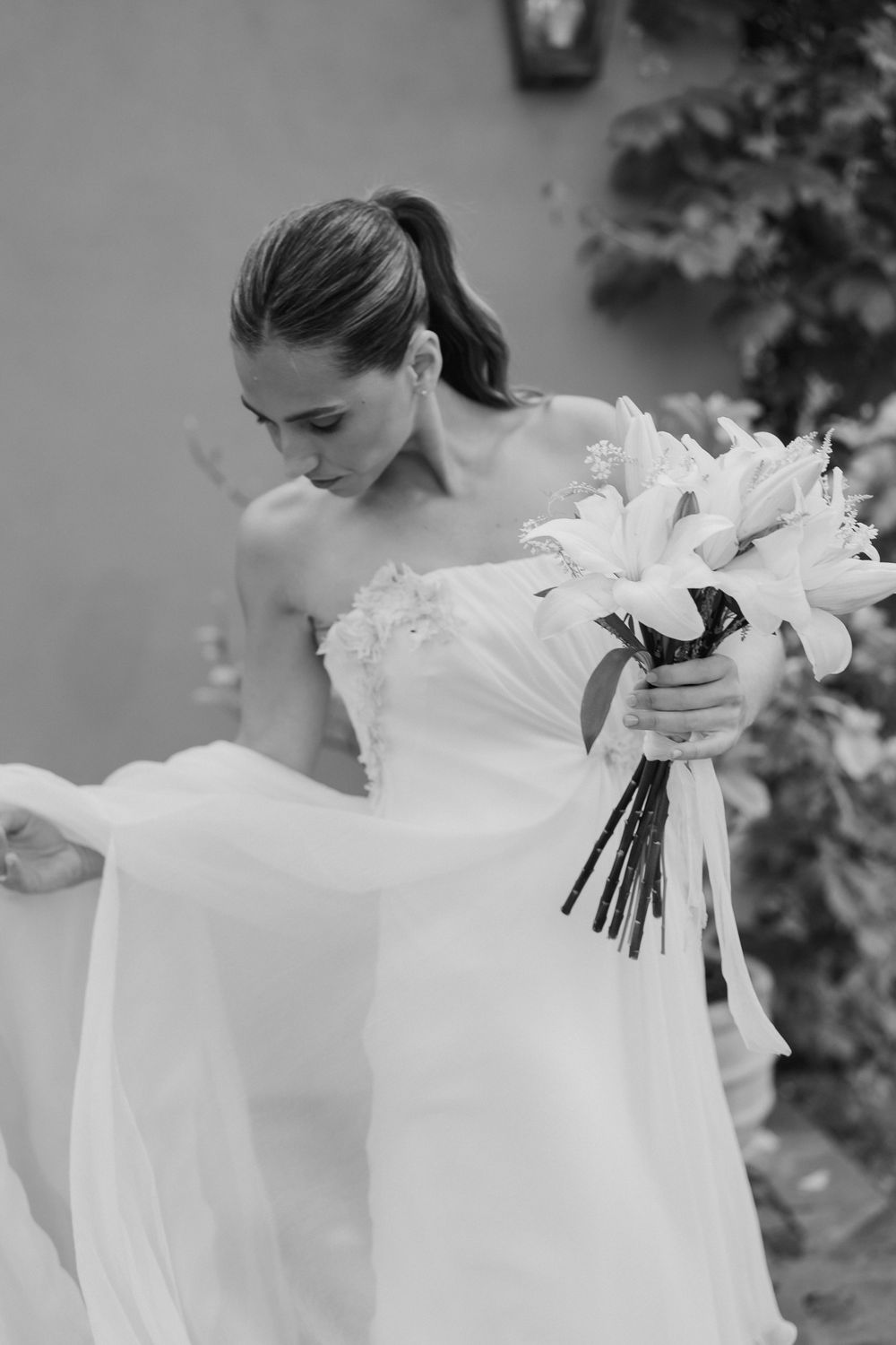 Black and white photo of a bride in a strapless gown with a flowing veil, looking down while holding a bouquet of white lilies.