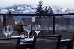 Wine glasses and a bottle on a balcony table overlooking snow-capped mountains and rooftops at dusk.