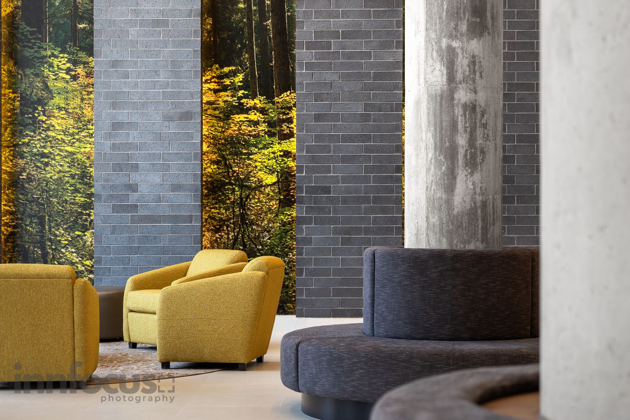 Modern lobby with yellow armchairs, gray brick columns and curved charcoal seating beside floor-to-ceiling windows overlooking autumn trees.