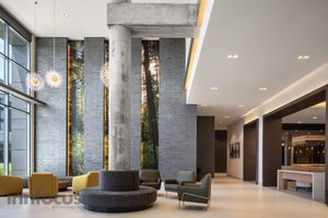 Modern building lobby with tall concrete columns, brick walls, forest-print panels, pendant lights, and yellow and gray lounge seating.