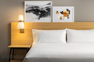 Modern hotel bed with crisp white linens, a wood headboard, wall-mounted lamp, bedside table and two framed abstract art prints above.