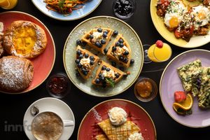 Overhead view of a brunch spread with blueberry toasts, pastries, fried eggs, a cappuccino, waffles with cream, avocado salad and juices.