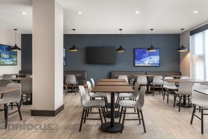 Modern hotel breakfast area with wooden tables, gray chairs, pendant lights, wall-mounted TVs and a blue accent wall.
