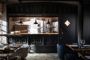 Modern restaurant interior with dark paneled walls, an open kitchen where a chef works, pendant lamps, and set tables with blue chairs.