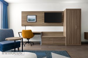 Modern hotel room with a wall-mounted TV, wood desk, yellow swivel chair, tall wardrobe, blue armchair, and patterned area rug.