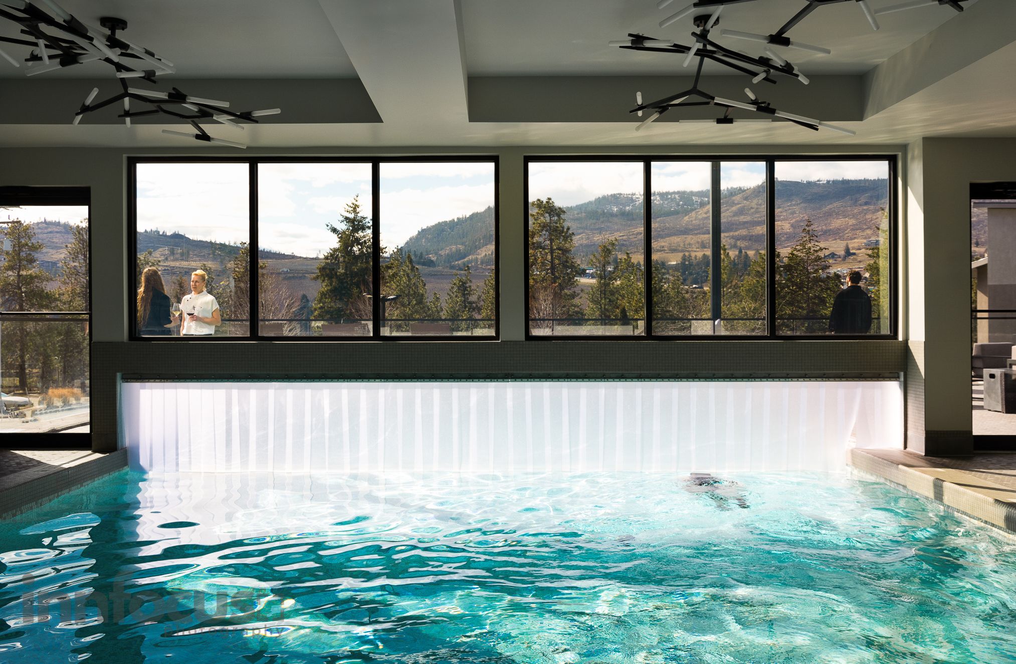 Indoor swimming pool with a swimmer, floor-to-ceiling windows and guests enjoying scenic mountain views at a luxury resort spa.