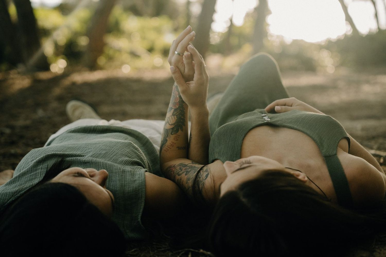 Two people with tattoos lie side by side on the forest floor, holding hands and gazing upward in a relaxed, sunlit woodland setting.