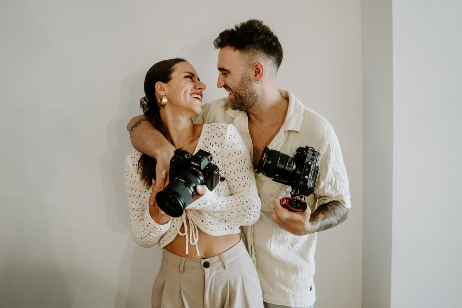 Smiling couple in neutral, cream-toned outfits embrace while each holding a professional camera against a plain light wall.