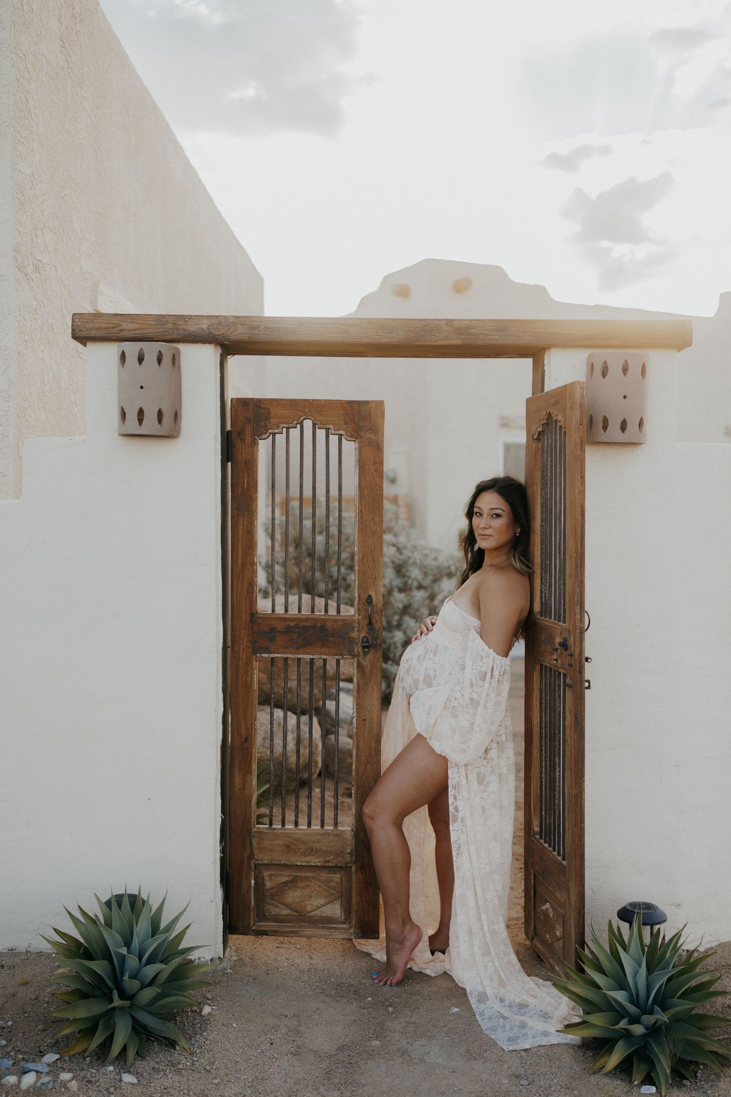 Pregnant woman in white lace gown poses at rustic wooden gate of Southwest adobe home.