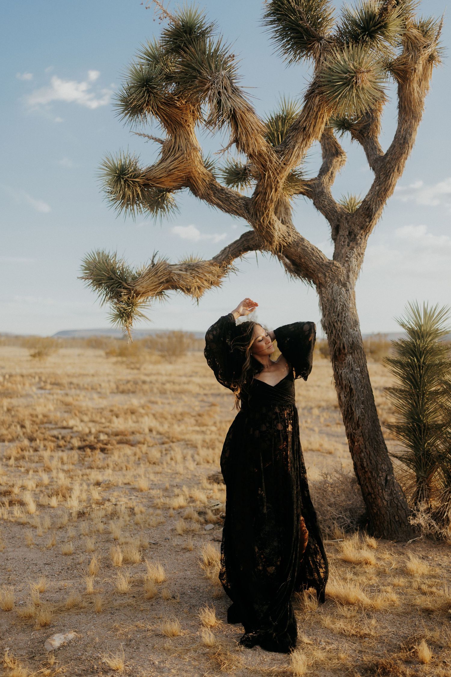 Woman in black dress poses by Joshua tree in desert landscape under blue sky.