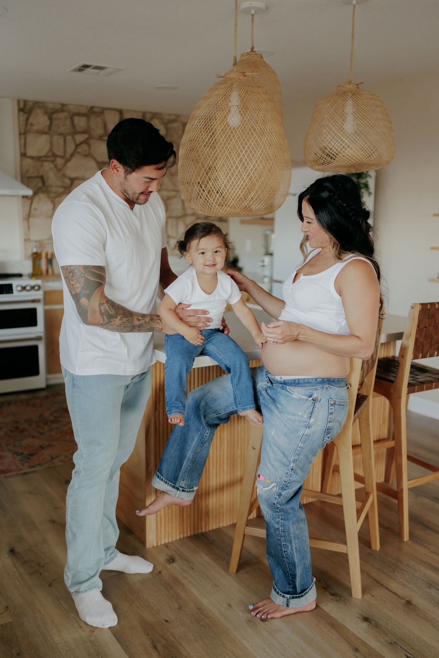Pregnant woman in white top and jeans stands with tattooed man and toddler in bright modern kitchen.