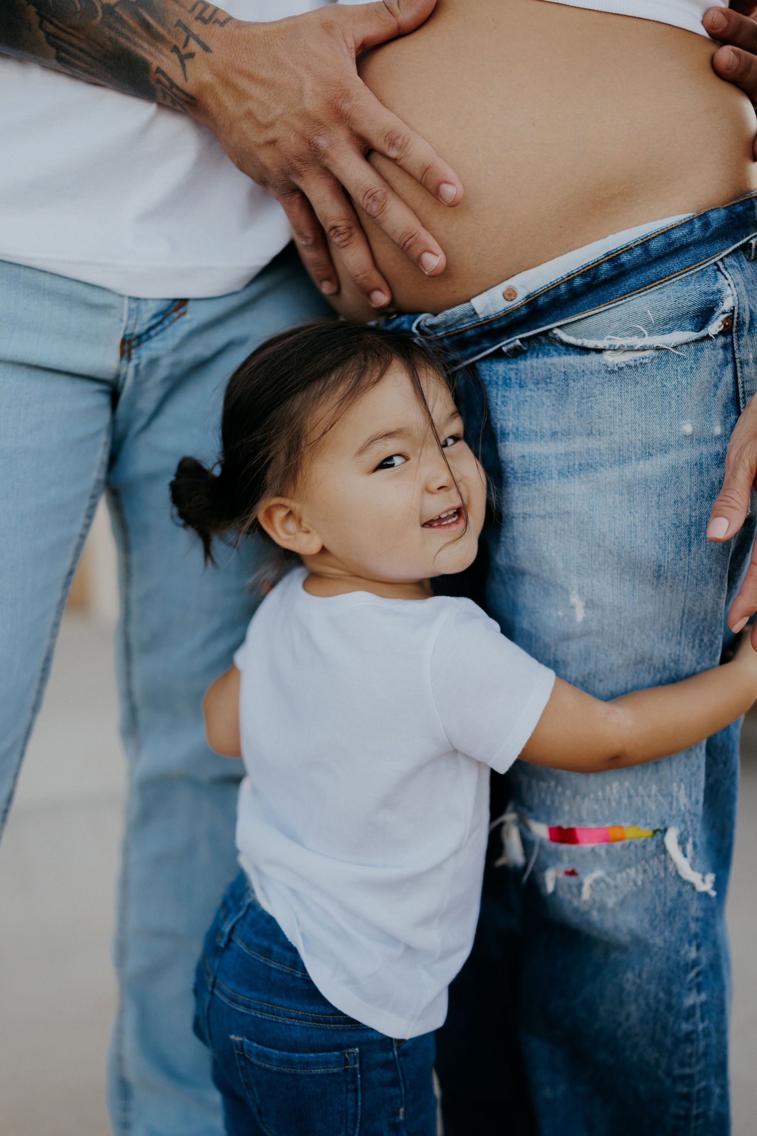 A young toddler girl hugs a pregnant woman's belly while standing between two adults in jeans outdoors.