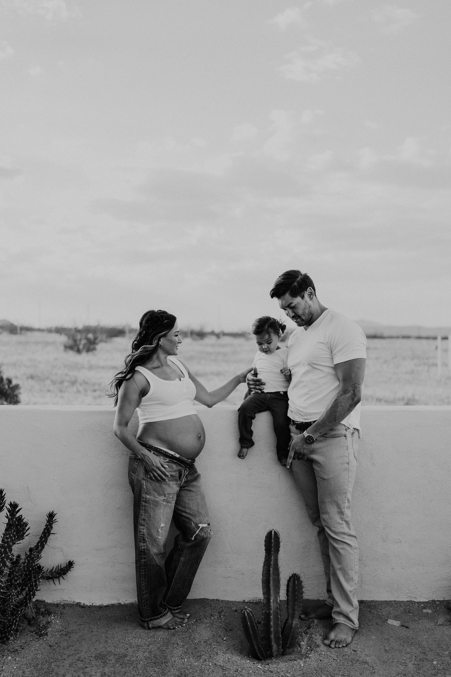 Black and white photo of pregnant woman and man with toddler by a wall with cacti outdoors.