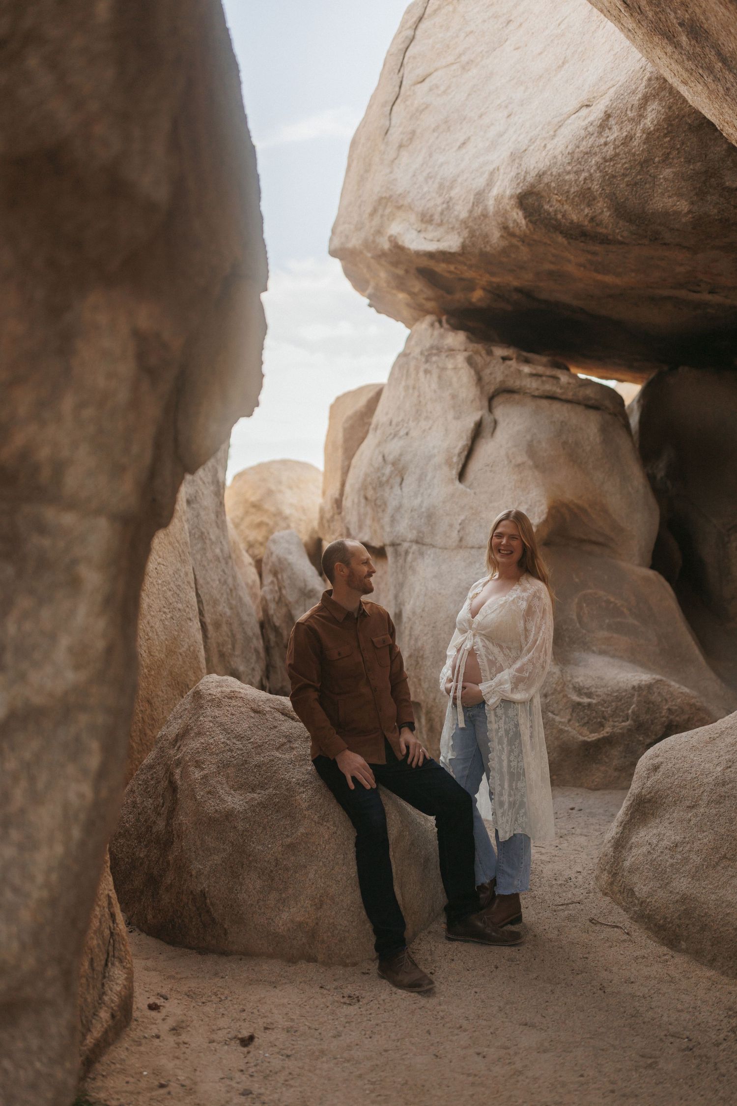 Couple posing among large boulders in a desert canyon, woman in white lace dress showing baby bump.