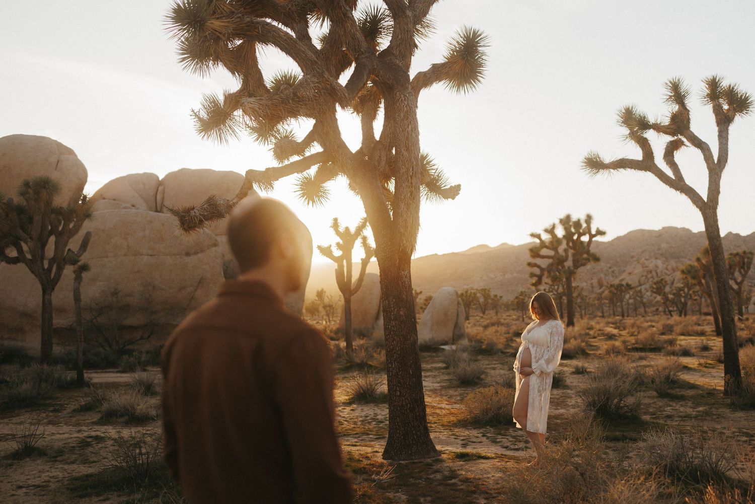 Two people stand among Joshua trees in golden desert sunset light, one in brown hoodie, one in white dress.