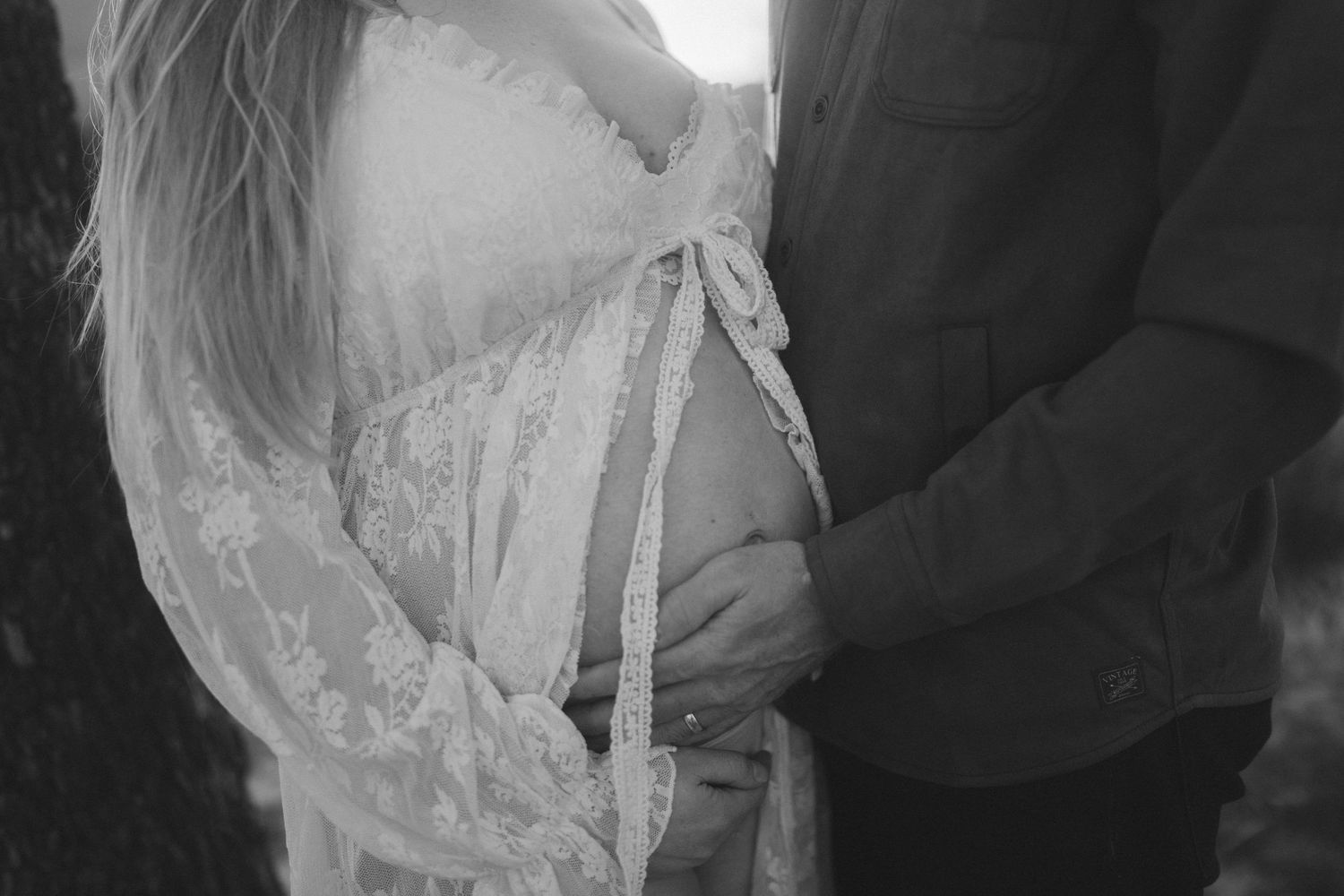 Black and white maternity photo of pregnant woman in lace dress with partner touching baby bump outdoors.
