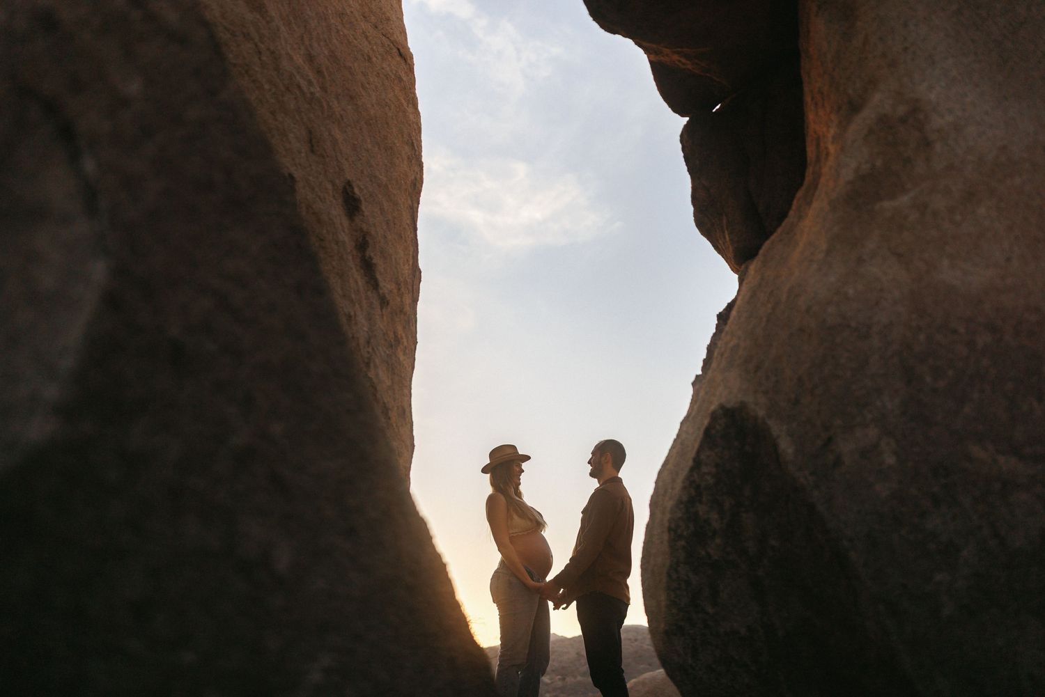 Couple holding hands silhouetted between large boulders at sunset in a desert landscape.