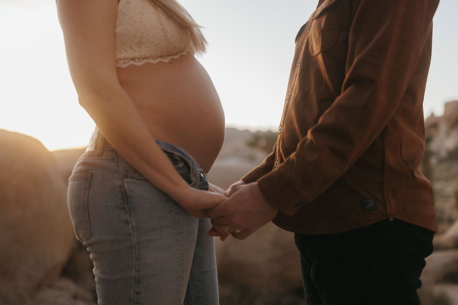 Pregnant couple holding hands outdoors at sunset, showing baby bump in jeans and brown jacket.