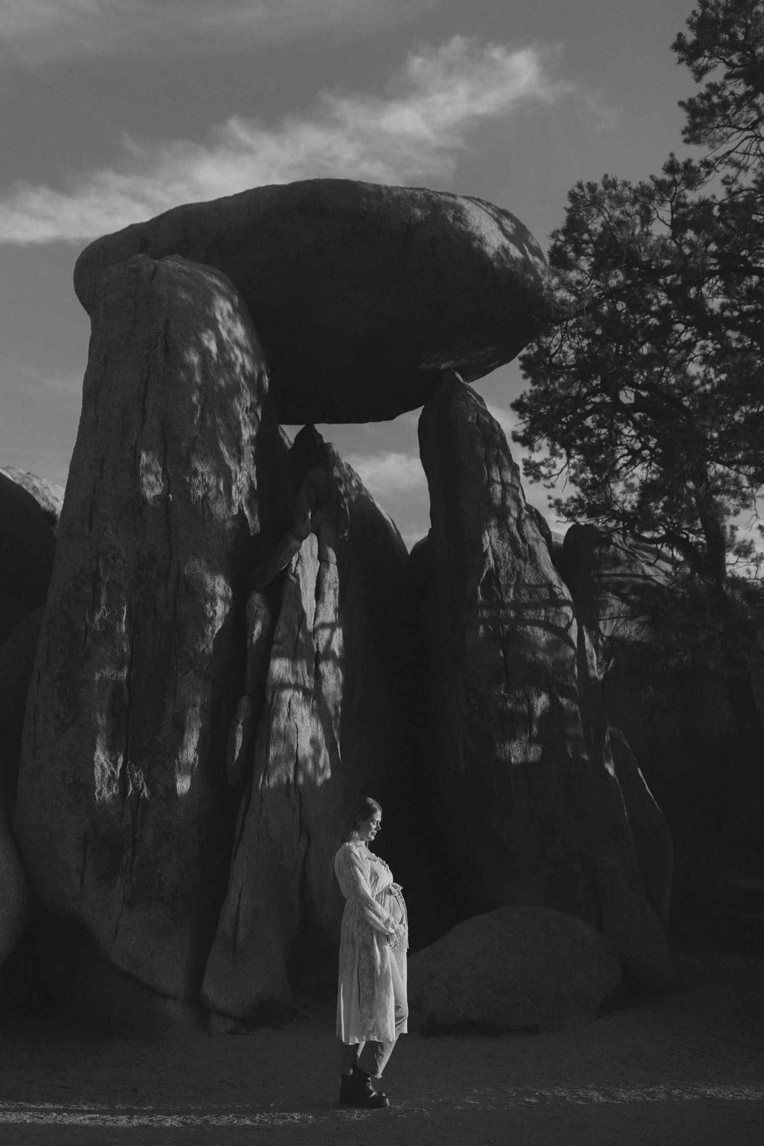 Black and white photo of a person in white coat standing beneath massive balanced rock formation resembling a dolmen.