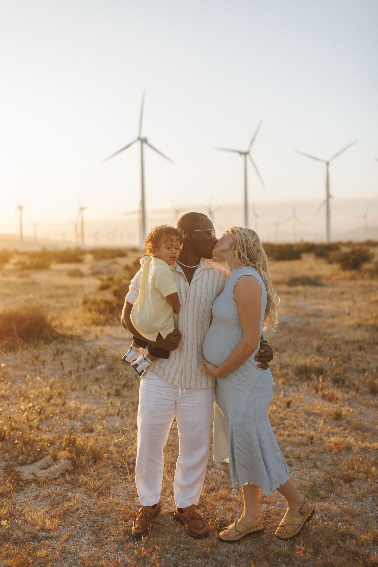 Interracial family with pregnant woman, man holding toddler, kissing at sunset near wind turbines in a golden field.