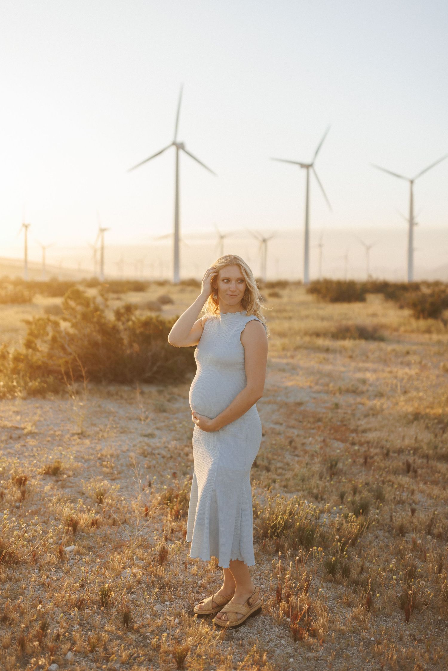 Pregnant woman in light blue dress standing in golden sunset field with wind turbines in background.
