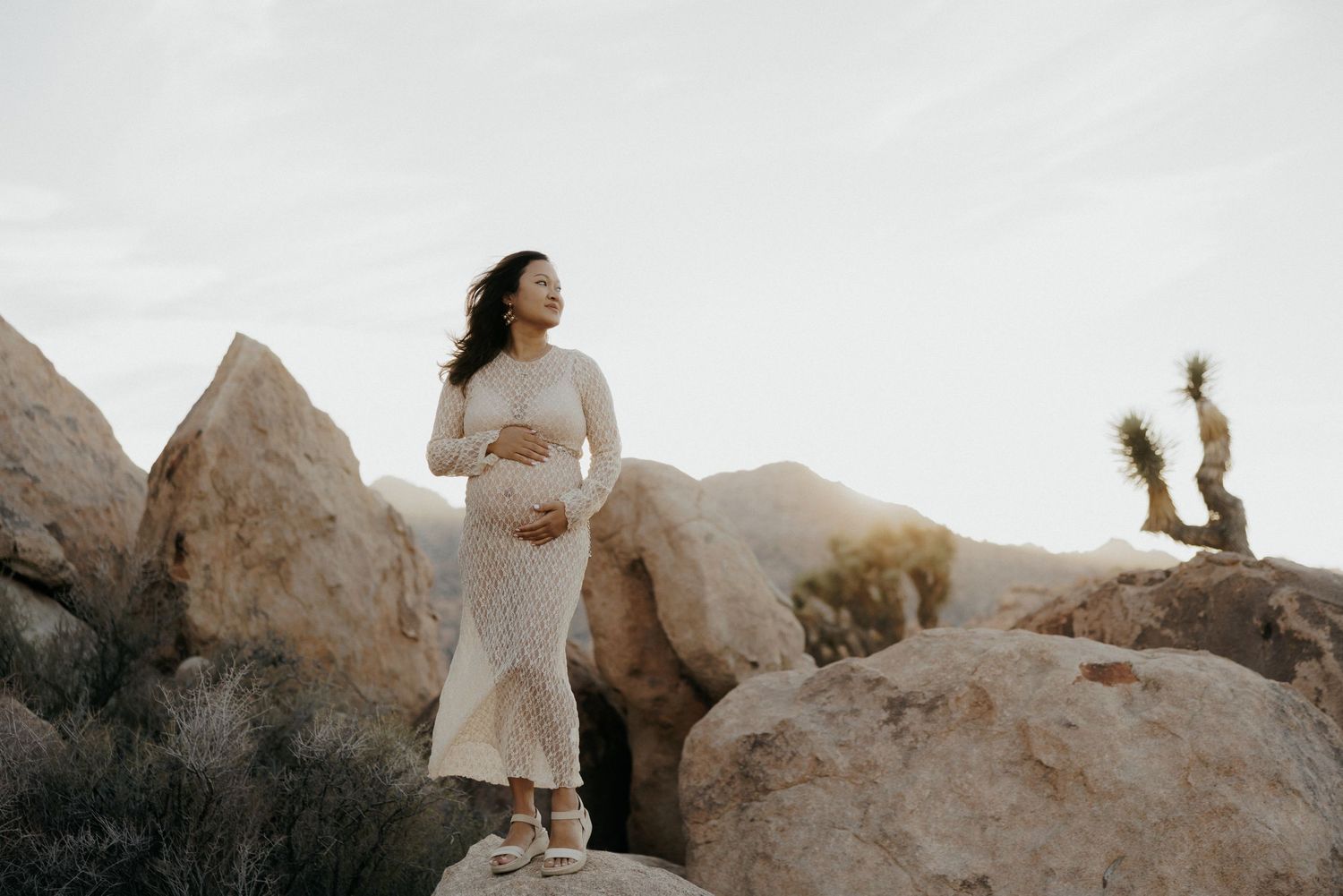 Pregnant woman in white lace dress standing on rocks in desert landscape at golden hour.