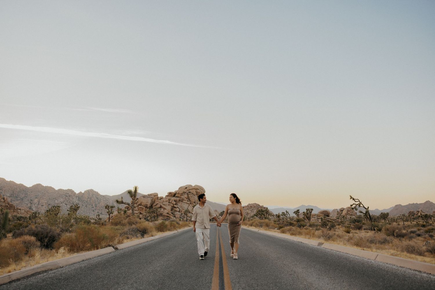 Couple holding hands walking down a desert road at dusk with Joshua trees and rocky mountains in the background.