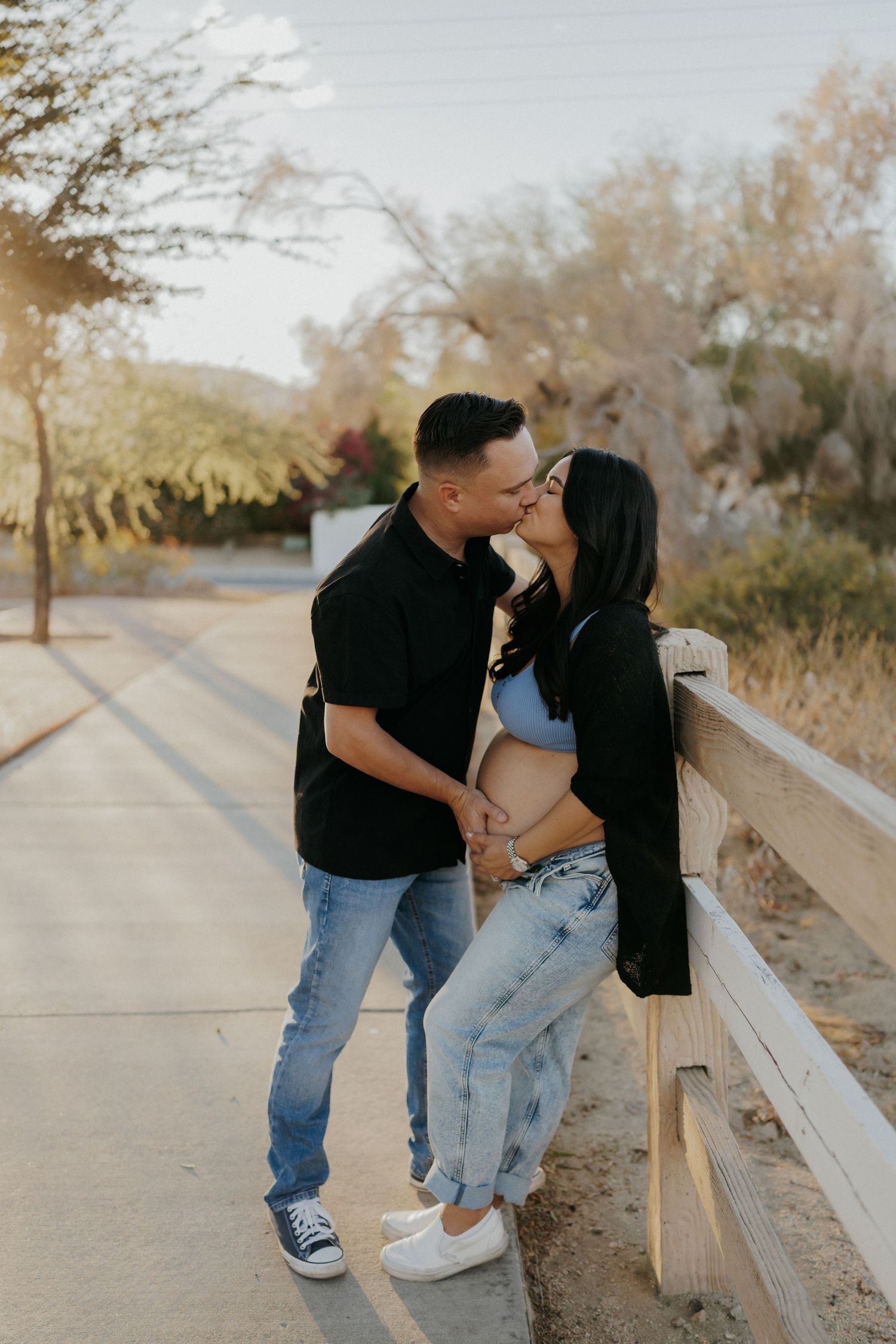 Couple sharing a kiss during maternity photo shoot outdoors, man tenderly holding pregnant woman's belly near wooden fence.