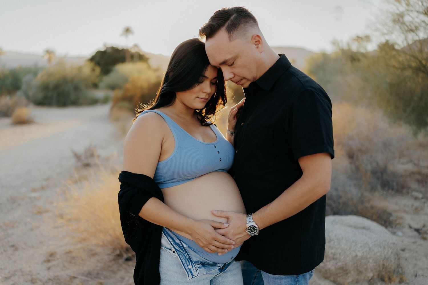 Couple embracing outdoors during maternity photo shoot, man holding pregnant woman's belly in desert landscape.