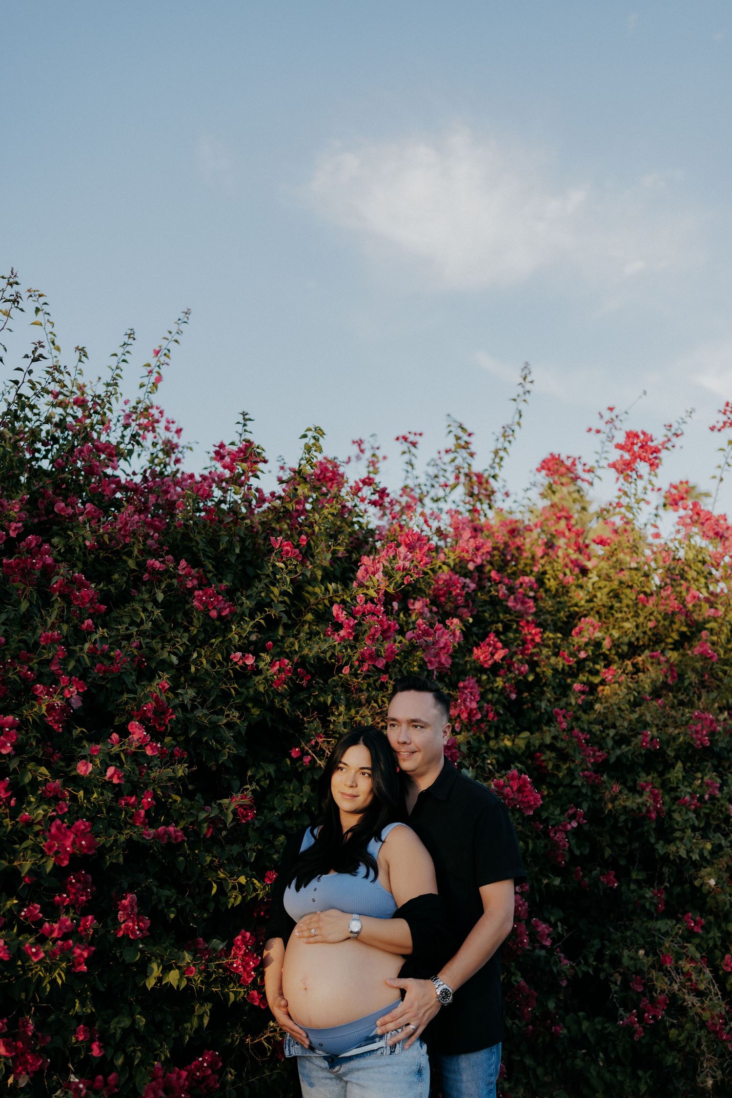 Couple posing for maternity photo in front of vibrant pink bougainvillea bushes under a blue sky.